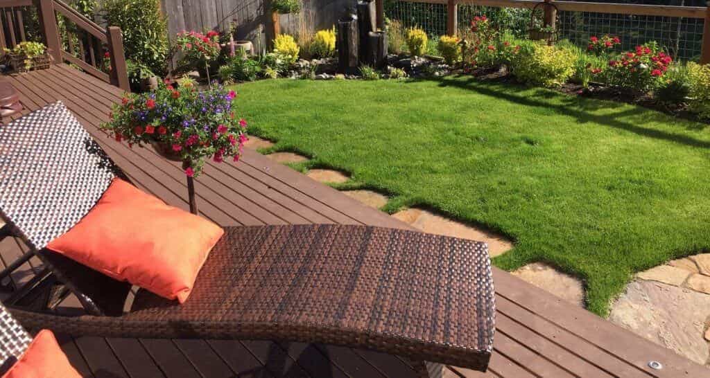 Wicker lounge chairs with orange cushions on a wooden deck overlook a lush green lawn bordered by colorful flowers and plants in a backyard garden. A hanging basket of flowers adds decoration near the chairs.
