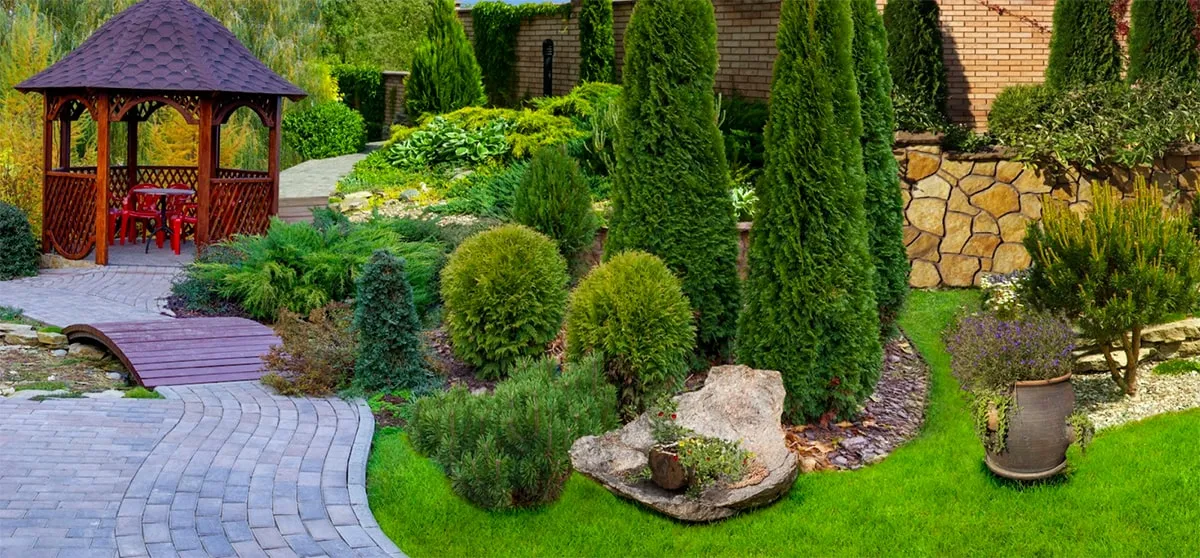 A landscaped garden with green shrubs, tall evergreen trees, a stone pathway, a wooden gazebo with red chairs, and a decorative stone and potted plants on the lawn.