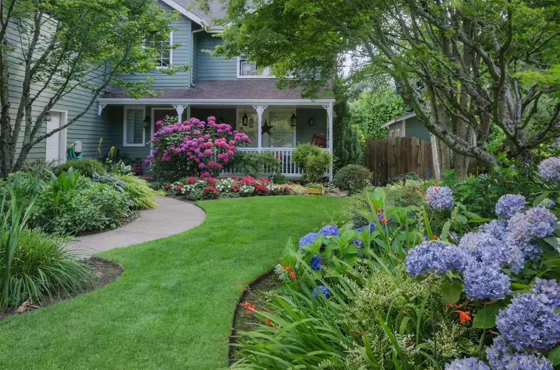 A lush garden with blooming flowers, manicured green lawn, and a curved path leads to a blue house with a white porch, surrounded by trees and shrubbery on a sunny day.