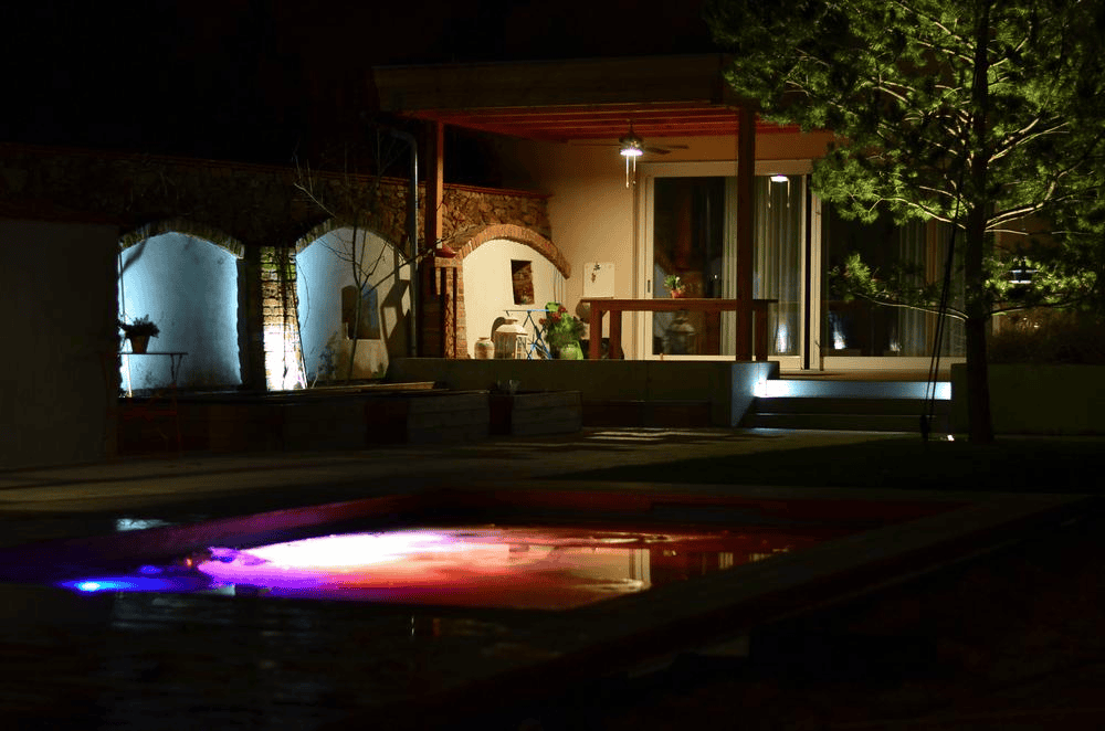 A cozy backyard at night features a glowing pool with colorful lights, a tree, and a well-lit patio attached to a house with large glass doors and potted plants.