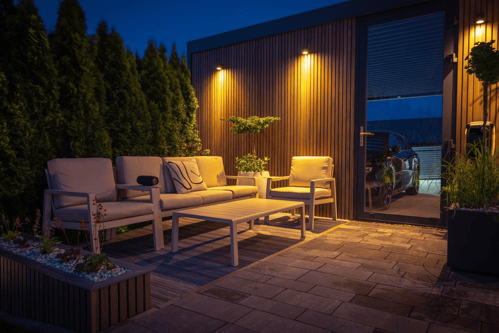 An outdoor patio with cushioned chairs, a sofa, and a coffee table is warmly lit by wall lights. The area is bordered by tall shrubs and modern wooden paneling, creating a cozy, inviting atmosphere at dusk.