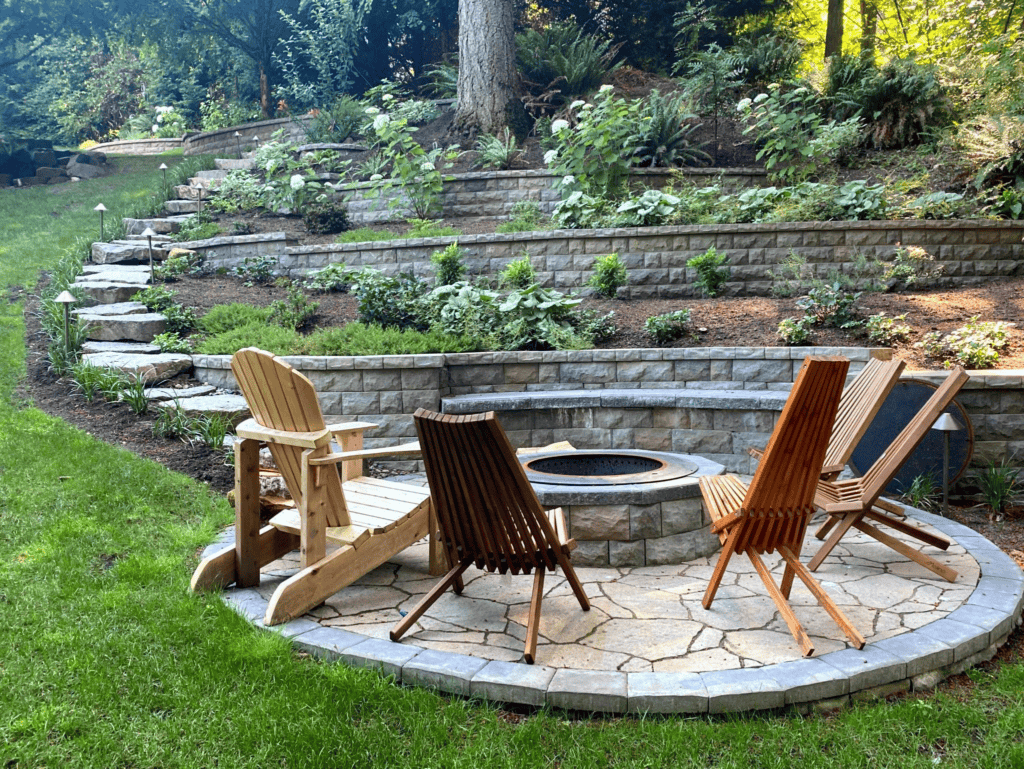 A circular stone patio with a fire pit is surrounded by four wooden chairs. The patio is set in a terraced garden with stone retaining walls, lush green plants, and a backdrop of tall trees.