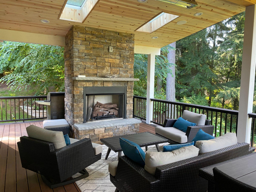 An outdoor covered patio with a stone fireplace, comfortable seating, a coffee table, and a grill. Wood-paneled ceiling with skylights.