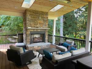 An outdoor covered patio with a stone fireplace, comfortable seating, a coffee table, and a grill. Wood-paneled ceiling with skylights.