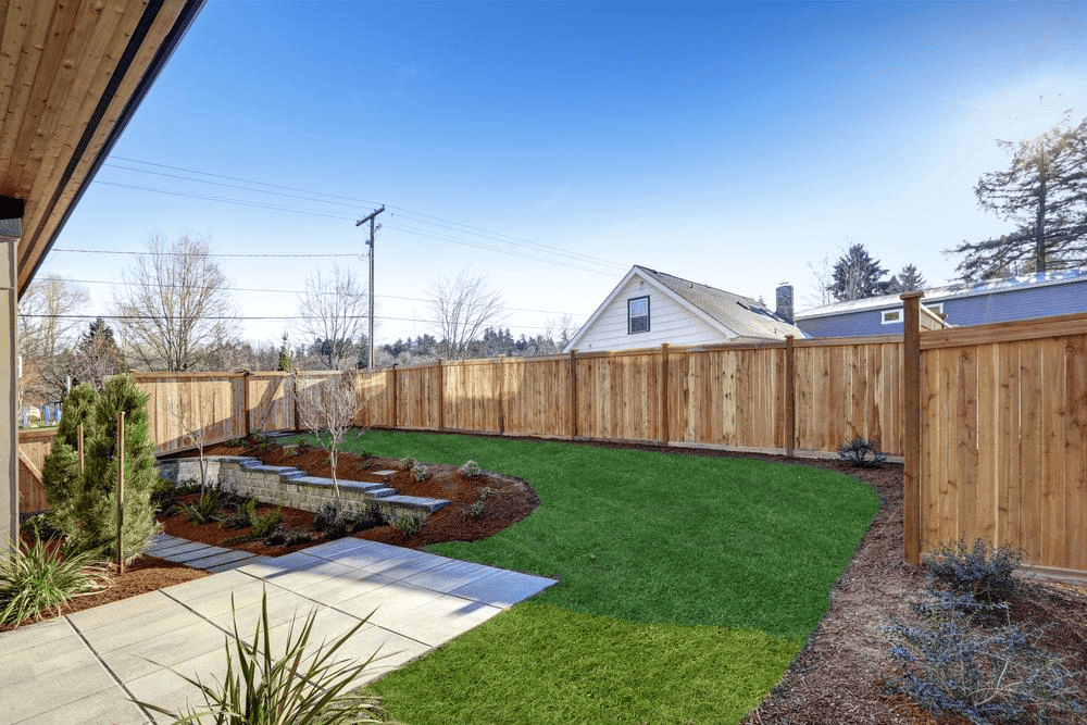 A neatly landscaped backyard with green grass, a paved patio area, small garden beds, and a tall wooden fence. A house and trees are visible outside the fence under a blue sky.