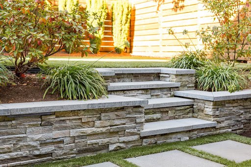 Three stone steps lead up between two raised garden beds with green plants, bordered by stacked stone walls. A wooden fence and greenery are visible in the background.
