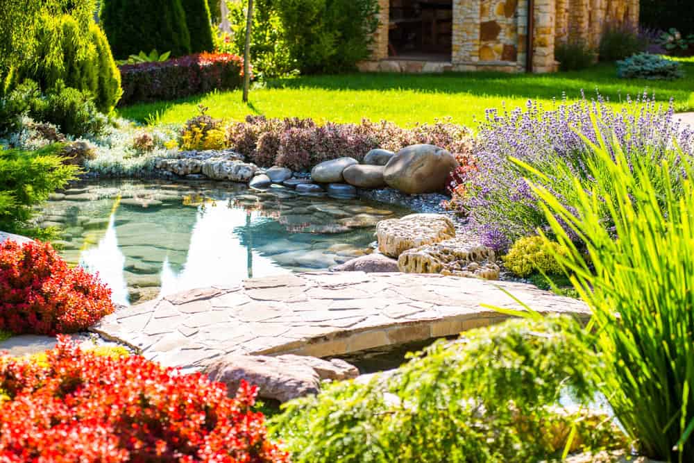 A landscaped garden with a small pond, stone bridge, colorful flowers, bushes, and green grass in bright sunlight.